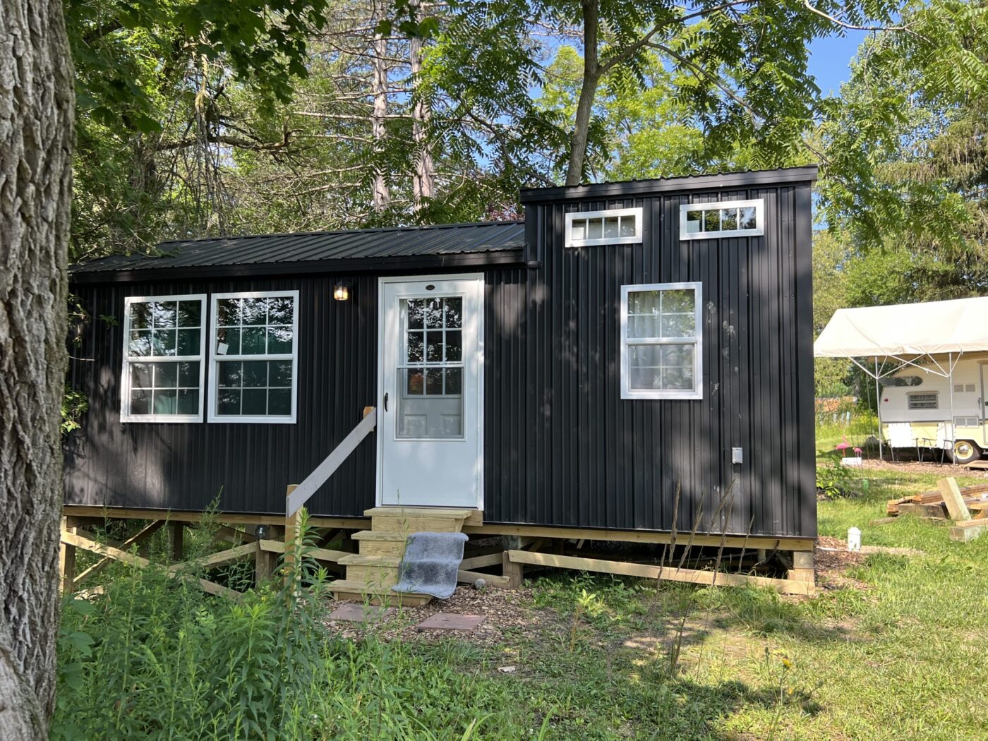 A sleek 12x18 cabin with a modern black metal exterior is nestled in a lush green setting. Large windows on the front and side allow ample natural light to flood the interior, while the white door with a glass panel adds a touch of elegance. Elevated on a wooden foundation, the cabin includes a small set of stairs leading to the entrance. Surrounded by trees and foliage, this cabin seamlessly blends into its natural environment, offering a tranquil and stylish retreat for various uses.