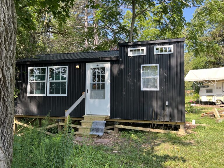 A sleek 12x18 cabin with a modern black metal exterior is nestled in a lush green setting. Large windows on the front and side allow ample natural light to flood the interior, while the white door with a glass panel adds a touch of elegance. Elevated on a wooden foundation, the cabin includes a small set of stairs leading to the entrance. Surrounded by trees and foliage, this cabin seamlessly blends into its natural environment, offering a tranquil and stylish retreat for various uses.