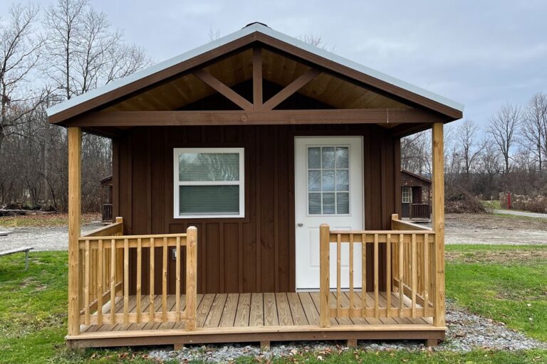 A quaint and compact wooden cabin featuring dark brown siding and a light-colored pitched metal roof. The front showcases a small, inviting covered porch with natural wooden railings and beams, adding rustic charm. The white-framed front door and adjacent window provide a clean contrast to the cabin’s dark exterior. The cabin is set on a simple gravel foundation, surrounded by green grass and a backdrop of bare trees, creating a peaceful and minimalist aesthetic suitable for cozy living or a weekend retreat.