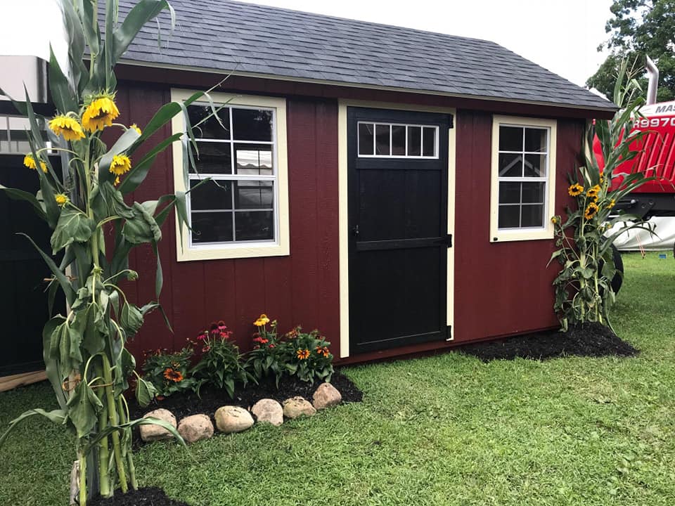 One of our garden sheds near Mt Morris NY - Dark red with black door white framed windows and black roof-green grass and sunflowers on each side
