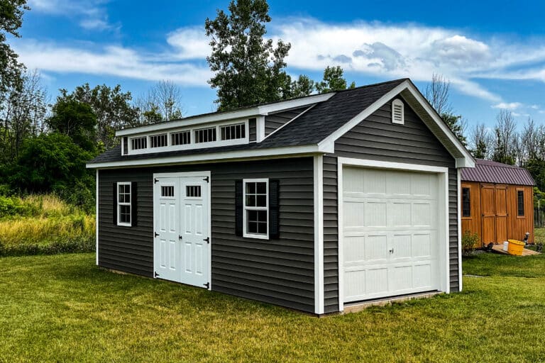 Dark gray prefab garage with white trim, a side door, and a roll-up garage door on a grassy lot.